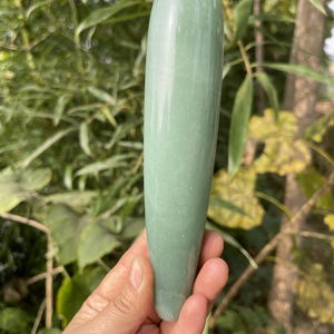 May include: A smooth, light green crystal wand, held in a hand. The wand is cylindrical with a rounded tip and a slightly tapered base. The background is blurred green foliage.
