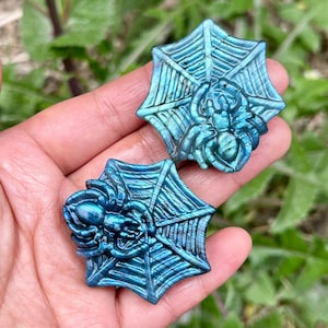 May include: Two blue-green carved gemstone pendants. Each pendant features a spider on a web design. The spider and web are intricately carved. The pendants are held in a hand against a background of green foliage.