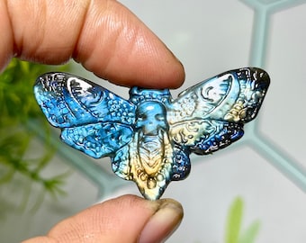 May include: A carved, iridescent moth pendant in shades of blue, teal, and gold. The wings feature intricate patterns and textures. The pendant is held between two fingers, showcasing its detailed craftsmanship.