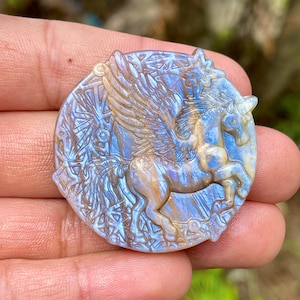 May include: A carved, round gemstone featuring a winged unicorn. The sculpture has a blue and gold iridescent color, with detailed wings and a flowing mane. The background has an etched design.