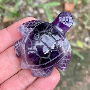 May include: A carved amethyst turtle figurine. The turtle is a deep purple color with a polished, glossy finish. The shell is segmented with a geometric pattern, and the head and flippers are detailed with carved lines.