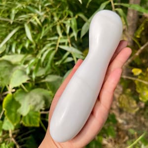 May include: A white, elongated massage tool made of stone, with a rounded top and a tapered handle. The tool is held in a hand against a backdrop of green foliage. The stone appears polished.