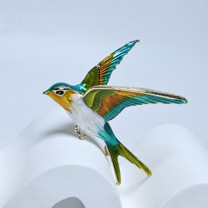 Puede incluir: Broche de pájaro colorido con las alas extendidas. El broche presenta un diseño detallado con una paleta de colores amarillo, blanco, turquesa, verde y dorado. Las alas del pájaro están extendidas y el broche tiene un alfiler dorado en la parte posterior.