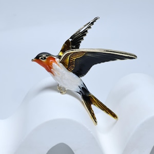 Puede incluir: Broche detallado con forma de golondrina en vuelo. El ave presenta cabeza negra, pecho naranja, vientre blanco y alas negras y doradas. El broche tiene detalles en color dorado.