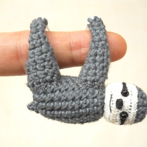 May include: A small grey crocheted sloth hanging from a finger. The sloth has white and black facial features.