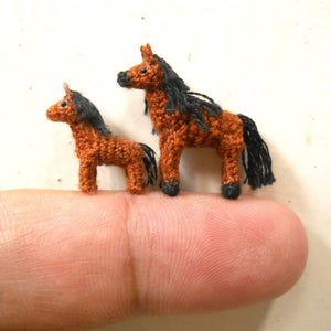 Op de afbeelding: Twee gehaakte paarden, een bruin en zwart, de andere kleiner en bruin met een zwarte manen en staart, zitten op een vinger.