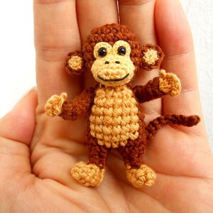 May include: A small crocheted monkey toy with brown fur, a yellow face, and black eyes. The monkey is sitting on an open hand.