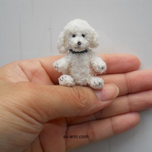 Puede incluir: Un pequeño juguete de caniche blanco de crochet con ojos negros y un collar negro. El juguete está sentado en la palma de una mano.