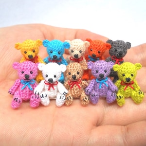 Puede incluir: Una colección de diez pequeños osos de peluche tejidos a crochet en varios colores, incluyendo rosa, morado, verde, amarillo, azul, blanco y gris. Los osos están todos sentados en una mano.
