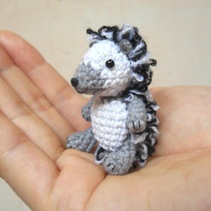Puede incluir: Un pequeño erizo de crochet con pelo blanco y gris y detalles negros. El erizo está sentado en una mano.
