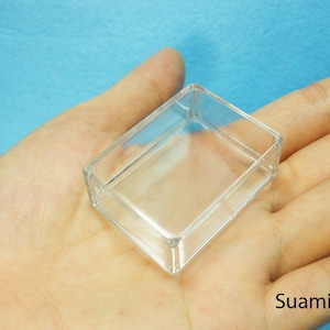 Może przedstawiać: Małe, przezroczyste prostokątne plastikowe pudełko z żebrowanym wzorem po bokach. Pudełko jest trzymane w dłoni osoby na niebieskim tle. Słowo "Suami" jest widoczne w prawym dolnym rogu.