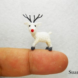 May include: A tiny white crocheted reindeer with black antlers and a red nose. The reindeer is standing on a finger.