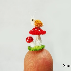 May include: A miniature crochet scene featuring a snail atop a red mushroom with white spots, next to a smaller mushroom. The snail has a yellow shell and white body. The mushrooms sit on a green base. The scene is displayed on a fingertip.