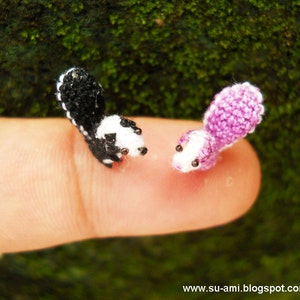 Puede incluir: Dos pequeños animales de crochet, uno blanco y negro y otro morado, están posados ​​en un dedo. El animal blanco y negro tiene una cola larga y una cabeza pequeña y redonda. El animal morado tiene una cola más corta y una cabeza redonda.
