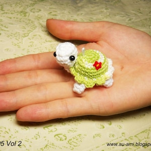 Puede incluir: Una pequeña tortuga de crochet con un cuerpo blanco y un caparazón verde. El caparazón tiene una flor roja en la parte superior. La tortuga está sentada en la mano de una persona.