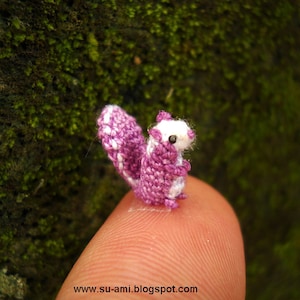 Puede incluir: Una pequeña figurita de ardilla de crochet de color morado y blanco. La ardilla está sentada en un dedo.