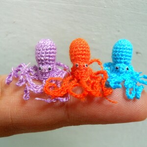May include: Three crocheted octopus figurines in purple, orange, and blue. The octopus are sitting on a finger.