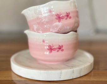 May include: Two stacked, pink and white ceramic teacups with floral designs and a matching marble coaster. The teacups have a gradient color scheme, transitioning from pink to white. The floral design features pink blossoms.
