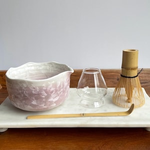 May include: A matcha tea set on a white marble tray. The set includes a light pink and white ceramic bowl, a clear glass cup, a bamboo whisk, and a bamboo scoop. The bowl has a textured, floral-like pattern.