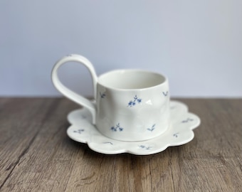 May include: A white ceramic teacup and saucer set. The cup has a unique handle and is decorated with small blue floral patterns. The saucer has a scalloped edge and matches the cup's design. The set is on a wooden surface.