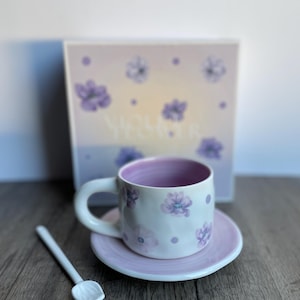 May include: A white ceramic teacup and saucer set with purple floral designs and a matching spoon. The interior of the cup is purple. A box with floral designs is in the background. The set is on a wooden surface.