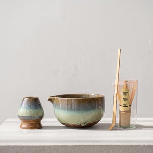May include: A matcha tea set featuring a ceramic whisk holder, a ceramic pouring bowl with a blue and green reactive glaze, a bamboo tea scoop, and a bamboo whisk inside a clear plastic container with a label reading 百本立 in black text.