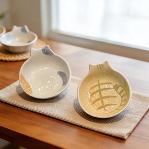 May include: Two ceramic cat-shaped bowls with a cream-colored interior and a light brown exterior. One bowl has a gray and tan design, while the other has a green striped design. The bowls are on a beige placemat.