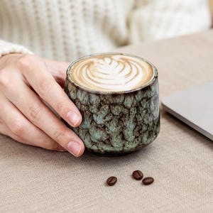 May include: A ceramic coffee cup with a textured, mottled green and brown exterior, filled with a latte featuring a decorative foam design. Three coffee beans are scattered on the surface.