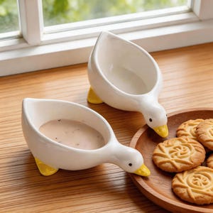 Op de afbeelding: Twee witte keramische ganzenvormige kommen met gele poten en snavels, elk met een lichtbruine vloeistof. De kommen staan op een houten oppervlak in de buurt van een houten bord met koekjes. De kommen staan in de buurt van een raam.