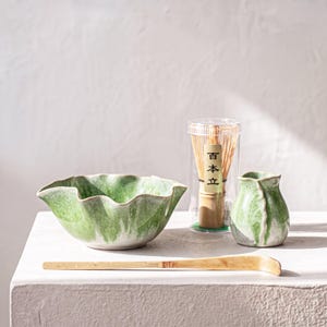May include: A green and white ceramic bowl with a wavy rim, a small matching ceramic pitcher, a bamboo whisk inside a clear plastic cylindrical case with the text 百本立, and a long, curved bamboo tea scoop arranged on a flat surface.