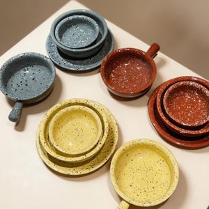 May include: A selection of speckled ceramic bowls and plates in various sizes and colours. The set includes bowls with handles, small bowls, and plates in shades of yellow, grey, and red. The speckled design adds a rustic touch to the tableware.