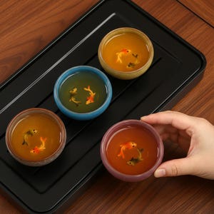 Puede incluir: Un juego de cuatro pequeñas tazas de té coloridas llenas de té, cada una con un diseño detallado de pez dorado en miniatura. Las tazas están dispuestas sobre una bandeja negra, con una mano parcialmente visible.