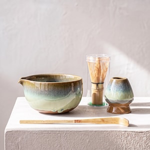 May include: A ceramic matcha bowl with a green and brown gradient glaze, a bamboo whisk inside a clear plastic cylindrical case, a ceramic whisk holder with a matching gradient glaze, and a long, curved bamboo tea scoop.