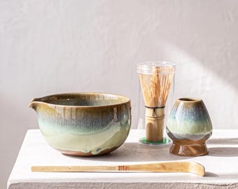 Może przedstawiać: Ceramiczna miseczka do matchy z zielono-brązowym szkliwem, bambusowa miotełka w przezroczystym plastikowym etui, ceramiczny stojak na miotełkę z pasującym szkliwem oraz długa, zakrzywiona bambusowa łyżeczka do herbaty.