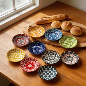 Puede incluir: Conjunto de pequeños platos decorativos de cerámica con diversos patrones coloridos. Los platos presentan diseños florales, geométricos y abstractos. Un pan y unos panecillos también están presentes sobre una superficie de madera.