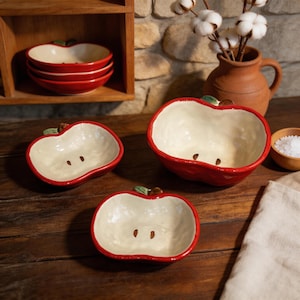 May include: A collection of ceramic apple-shaped bowls. The bowls are red with a cream-colored interior and brown seed details. A stack of bowls is on a shelf, and others are arranged on a wooden surface. A small vase with cotton stems is in the background.