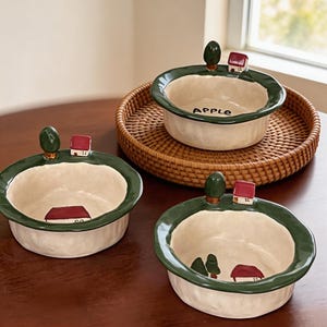 Könnte beinhalten: Drei Keramikschalen mit cremefarbenem Innenraum und dunkelgrünem Rand. Jede Schale hat ein Miniaturhaus- und Baumdesign. Eine Schale hat das Wort "APPLE" auf der Innenseite. Die Schalen stehen auf einem geflochtenen Tablett.