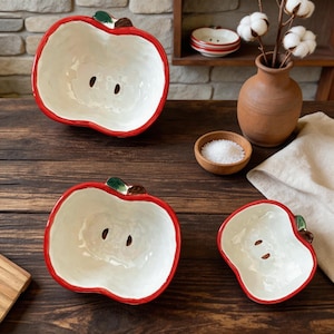 May include: A collection of ceramic apple-shaped bowls. The bowls have a white interior, a red rim, and a green and brown stem detail. The bowls vary in size and are arranged on a wooden surface.