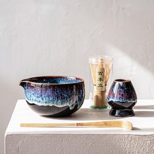 Puede incluir: Un cuenco de cerámica para matcha con esmalte de goteo azul oscuro y morado, un batidor de bambú en un estuche de plástico transparente con el texto 百本立, un soporte de cerámica a juego y una cuchara de té de bambú larga y curvada.
