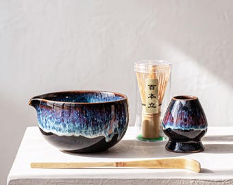 May include: A ceramic matcha bowl with a dark blue and purple drip glaze, a bamboo whisk inside a clear plastic case with the text 百本立, a matching ceramic whisk holder, and a long, curved bamboo tea scoop arranged on a flat surface.