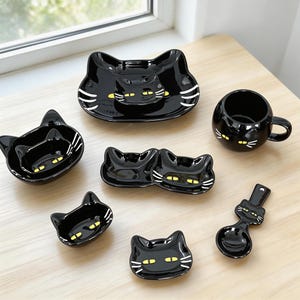 May include: A set of black ceramic tableware, each piece shaped like a cat's face. The collection includes plates, bowls, and a mug, with yellow eyes and white whisker details. The items are displayed on a light wooden surface.