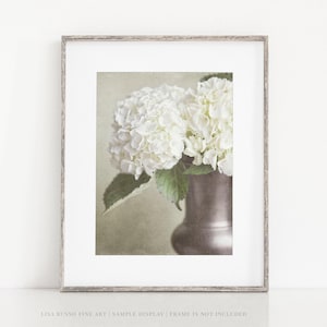 May include: A framed print featuring a still life of white hydrangeas in a metallic vase. The flowers are in full bloom, with lush green leaves. The vase is a muted silver color, and the background is a soft, neutral tone.