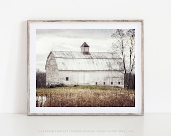 May include: Framed print of a white barn with a red cupola, set against a cloudy sky. The barn has a weathered appearance, with a metal roof and multiple windows. The foreground features tall, dried grasses and a body of water.