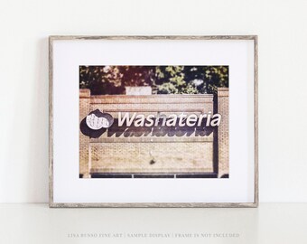 Op de afbeelding: Ingelijste print met een vintage bord "Washateria" in retrostijl. Het bord is wit met een gestileerd ontwerp, geplaatst tegen een bakstenen gebouw. De print zit in een houten lijst.
