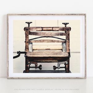 May include: A framed print featuring a vintage wooden washing machine. The antique laundry wringer has a light-colored roller and is set against a neutral background. The print is in a light-colored wooden frame.
