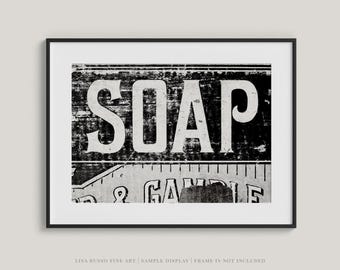 May include: Black and white framed print featuring the word "SOAP" in large, bold letters. The print has a distressed, vintage aesthetic, with visible texture and wear. The bottom of the print shows the words "& GAMBLE".