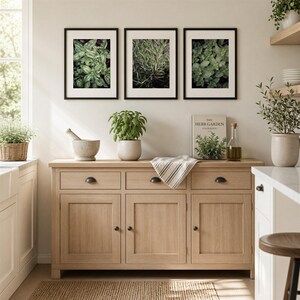May include: A light oak wooden sideboard with three drawers and three cabinets, featuring black hardware. Above it hang three framed botanical prints. On the sideboard are a mortar and pestle, a potted plant, a book, and a glass oil bottle.