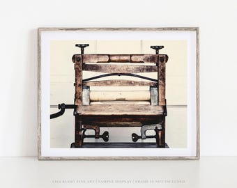 May include: A framed print of an antique wooden clothes wringer. The wringer has a light-colored roller and is made of weathered wood with metal hardware. The image is in a neutral color palette, with a cream-colored background.