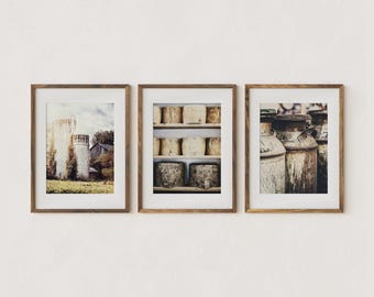 May include: Set of three framed prints featuring rustic farm scenes. One print shows a silo and barn, another displays aged cheese wheels, and the third depicts vintage milk cans. The prints have a warm, neutral color palette and are displayed in wooden frames.