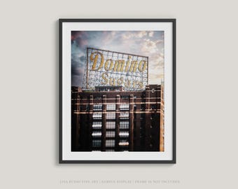 Op de afbeelding: Ingelijste kunstdruk met het Domino Sugar-bord op een bakstenen gebouw. Het bord heeft gouden letters en staat tegen een bewolkte lucht. Het gebouw heeft veel ramen.